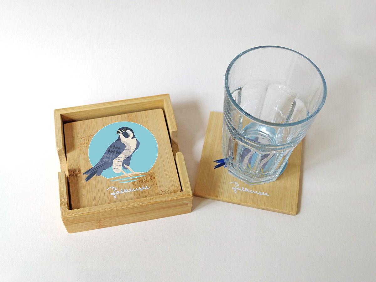 falkensee-logo-falke-untersetzer-isabel-gewecke Bambusuntersetzer mit Falkenillustration und Schriftzug „Falkenreise“ neben Glas – Design: Isabel Gewecke.