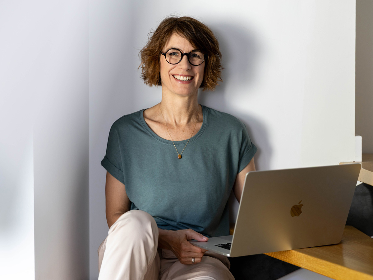 Frau mit Brille und grünem Shirt sitzt lächelnd mit Laptop auf dem Schoß auf einer Stufe – Foto: Tanja Marotzke.