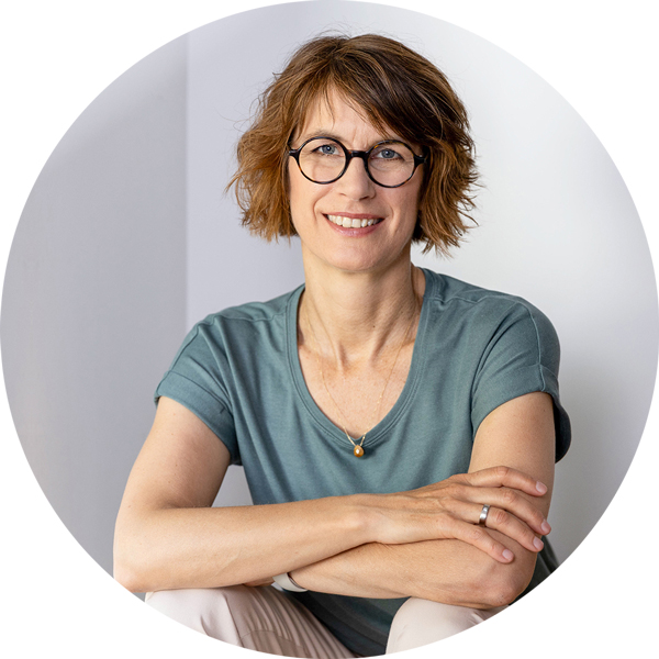 Frau mit kurzem, lockigem Haar sitzt locker mit gekreuzten Armen vor heller Wand – – Foto: Tanja Marotzke.