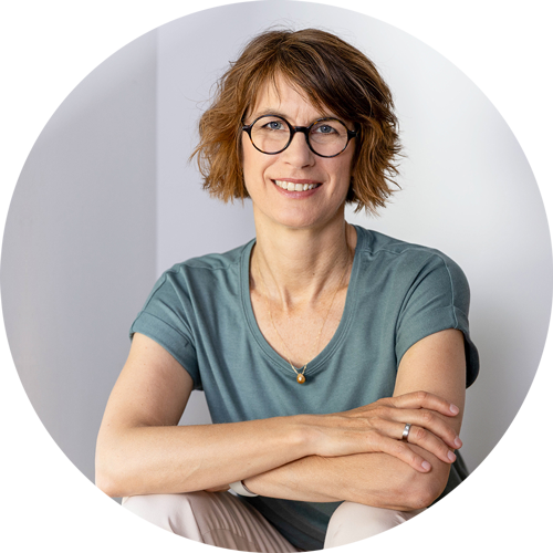 isabel-gewecke-portraet-rund Frau mit kurzem, lockigem Haar sitzt locker mit gekreuzten Armen vor heller Wand – – Foto: Tanja Marotzke.